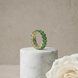 Könnte beinhalten: Ein goldener Eternity-Ring mit einer durchgehenden Reihe von oval geschliffenen grünen Smaragden in einer Krappenfassung. Der Ring steht aufrecht auf einem zylindrischen Sockel aus weißem Marmor und hebt die leuchtend grüne Farbe der Steine hervor.