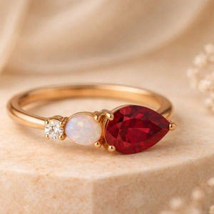 Könnte beinhalten: Ein Goldring mit einem birnenförmigen roten Edelstein, einem runden weißen Opal und einem kleinen runden klaren Diamanten. Die Steine sind in einer Reihe auf dem dünnen Goldband angeordnet und bilden ein Design, das den Kontrast der Elemente betont.