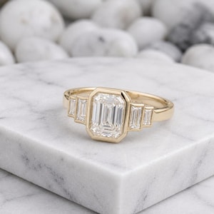 Könnte beinhalten: Ein Goldring mit einem zentralen Smaragdschliff-Stein in einer Zargenfassung, flankiert von zwei kleineren Baguette-Schliff-Steinen auf jeder Seite. Die Ringschiene ist glatt und poliert und zeigt ein klares, geometrisches Design mit facettierten Steinen.