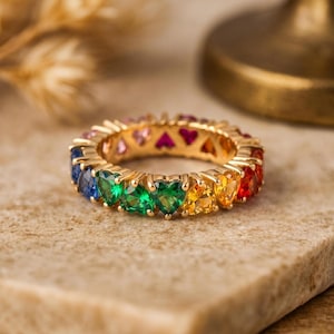 Könnte beinhalten: Ein goldener Eternity-Ring mit einer durchgehenden Reihe herzförmiger Edelsteine in einem lebendigen Regenbogenspektrum, darunter rote, orange, gelbe, grüne und blaue Steine, die in einen polierten Metallrahmen gefasst sind, der den Ring umschließt.