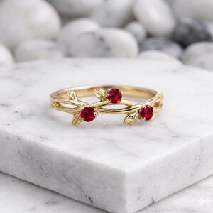Könnte beinhalten: Ein goldener Ring mit einem Band, das mit zarten Blattmotiven gestaltet ist und drei runde, leuchtend rote Rubine an der Oberseite aufweist. Das Metall hat eine polierte Oberfläche und die astartige Struktur verleiht dem Ring ein organisches Aussehen.