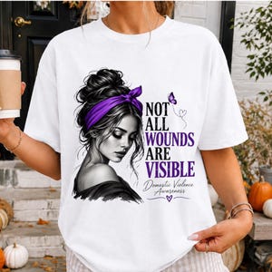 Può includere: Una t-shirt bianca con un'illustrazione in bianco e nero di una persona con una fascia viola e uno chignon spettinato. Il testo recita: NOT ALL WOUNDS ARE VISIBLE Domestic Violence Awareness, accompagnato da una farfalla viola e un cuore.