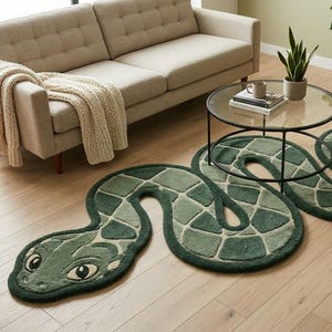 May include: A long, winding area rug shaped like a snake, featuring a segmented pattern in various shades of dark and light green. The rug has a detailed head with large, almond-shaped eyes and a textured, tufted surface that follows the serpentine form.