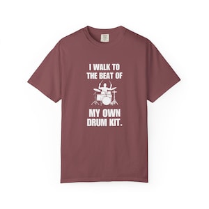 May include: A maroon short-sleeved t-shirt featuring a white graphic of a person playing drums. The text printed in white reads: I WALK TO THE BEAT OF MY OWN DRUM KIT. The shirt has a crew neckline and a standard fit, made of a solid-coloured fabric.
