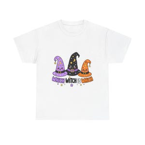 May include: A white t-shirt featuring a graphic of three cartoon witch hats in purple, black, and orange. Each hat has a smiling face, a belt, and star and moon patterns. Below the hats, the text reads "WHICH WITCH IS WHICH" in colourful letters.