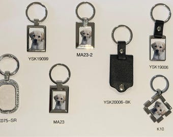 May include: A collection of metal keychains featuring various shapes including rectangular, oval with rhinestones, and square frames. Several keychains display a photograph of a yellow Labrador puppy. Labels include YSK19099, MA23-2, YSK20006-BK, YSK19006, SX075-SR, MA23, and K10.