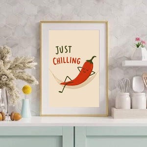 May include: A framed wall art print featuring a cartoon red chili pepper with a face, arms, and legs, relaxing in a hammock. The text "JUST CHILLING" is printed in dark green and red above the chili pepper on a light cream background.