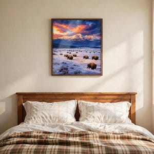 Buffalo wall art square framed print above a wooden bed in a cosy cabin bedroom, snowy mountain landscape with a bison herd at sunset