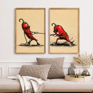 May include: Two framed wall art prints featuring a red chilli pepper character wearing sunglasses and trainers. In the left print, the chilli pulls a rope, and in the right print, the chilli pulls the other end of the rope with an angry expression.