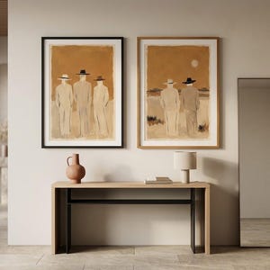 May include: Two framed wall art prints featuring figures in hats against a warm ochre background, displayed above a light wood console table with a terracotta vase, a small lamp, and a tall rectangular floor mirror standing to the right.