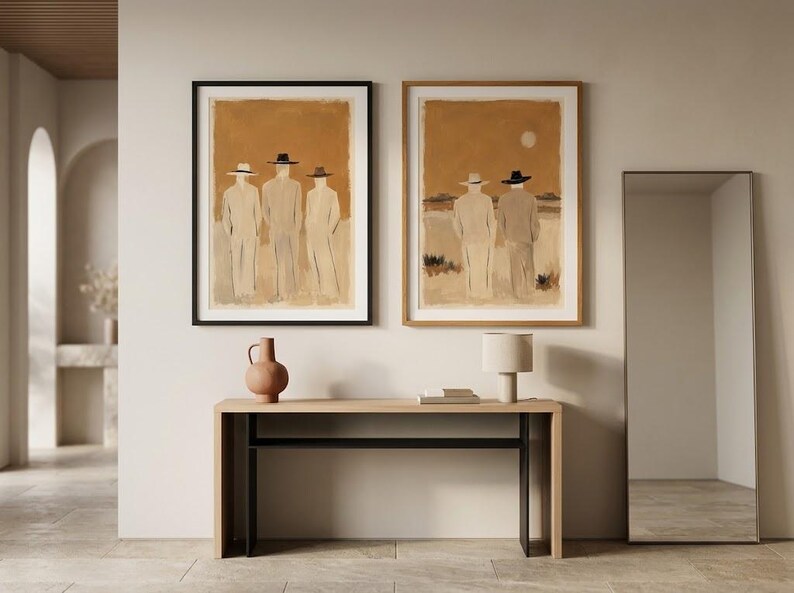 May include: Two framed wall art prints featuring figures in hats against a warm ochre background, displayed above a light wood console table with a terracotta vase, a small lamp, and a tall rectangular floor mirror standing to the right.