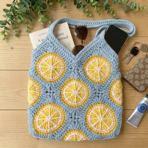 May include: A light blue crochet tote bag featuring a repeating pattern of yellow and white lemon slice motifs. The bag contains a book, sunglasses, and a smartphone. Nearby items include a set of keys, a patterned wallet, and a tube of paint.