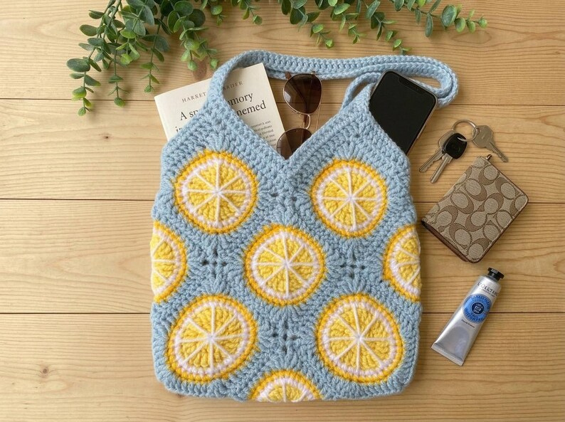 May include: A light blue crochet tote bag featuring a repeating pattern of yellow and white lemon slice motifs. The bag contains a book, sunglasses, and a smartphone. Nearby items include a set of keys, a patterned wallet, and a tube of paint.