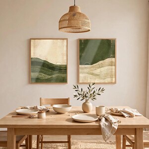 May include: Two rectangular wall art prints in light wood frames featuring abstract landscape designs with layered green and beige tones, geometric sun motifs, and thin line art elements displayed on a plain wall above a wooden dining table.