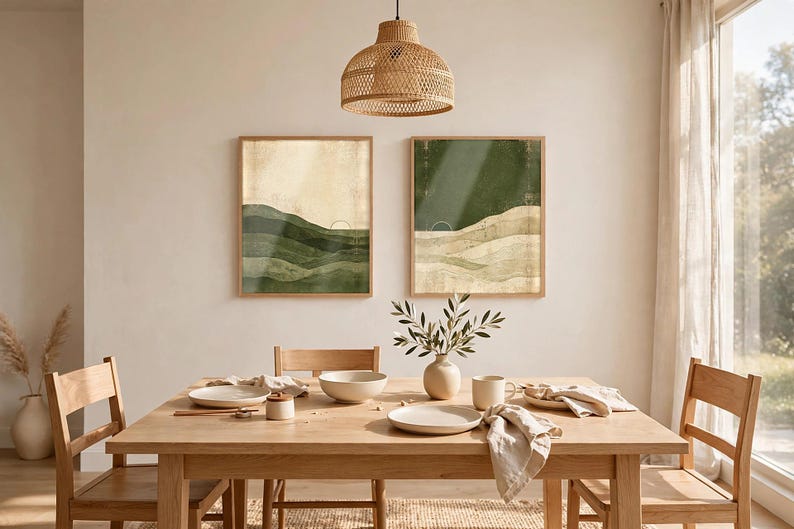 May include: Two rectangular wall art prints in light wood frames featuring abstract landscape designs with layered green and beige tones, geometric sun motifs, and thin line art elements displayed on a plain wall above a wooden dining table.
