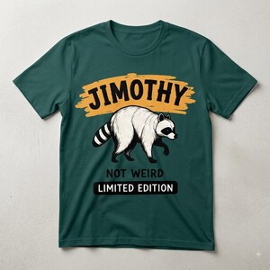 May include: A dark teal short-sleeve t-shirt featuring a graphic of a white raccoon with black markings. Above the raccoon is the text JIMOTHY in black on a yellow brushstroke background. Below the raccoon is the text NOT WEIRD LIMITED EDITION.