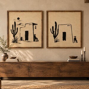 May include: Two square wall art pieces in dark wood frames featuring black ink illustrations on a beige background. The left print shows a desert building, a cactus, and a seated figure. The right print depicts a similar building and cactus.