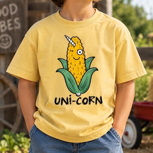 May include: A mustard yellow short-sleeve t-shirt featuring a graphic of a yellow corn cob with a white unicorn horn, a single eye, and green husks. Below the graphic, the text UNI-CORN is printed in a bold, black, hand-drawn style font.