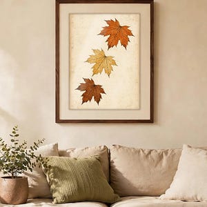 May include: A framed wall art print featuring three maple leaves in varying shades of orange, yellow, and brown, arranged vertically against a textured, off-white background. The artwork is enclosed in a dark brown wooden frame with a wide mat.