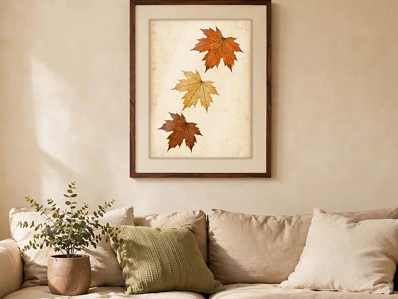 May include: A framed wall art print featuring three maple leaves in varying shades of orange, yellow, and brown, arranged vertically against a textured, off-white background. The artwork is enclosed in a dark brown wooden frame with a wide mat.