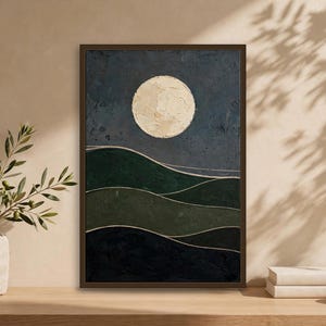 Japandi wall art print with a large cream full moon over dark green rolling hills, framed and leaning on a wooden surface with a stone vase and books — moody abstract landscape, digital download