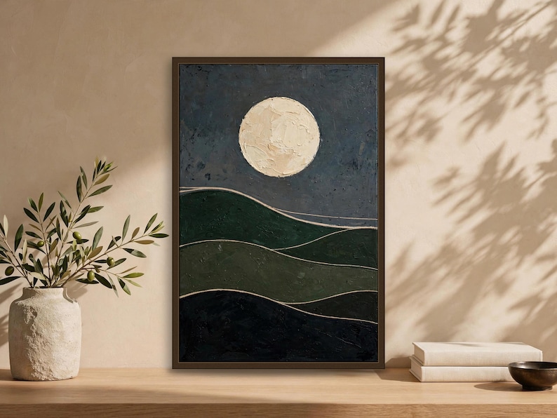 Japandi wall art print with a large cream full moon over dark green rolling hills, framed and leaning on a wooden surface with a stone vase and books — moody abstract landscape, digital download