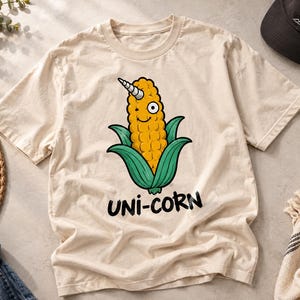 May include: A beige short-sleeve t-shirt featuring a graphic of a yellow corn cob with a white unicorn horn on its head, green husks, and a smiling face. Below the illustration, the text UNI-CORN is printed in bold, black, hand-drawn style letters.