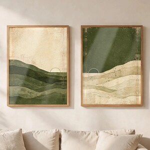 May include: Two framed wall art prints featuring abstract landscape designs. The left print shows green rolling hills against a cream sky, while the right print displays cream-colored dunes under a dark green sky, both with a thin sun arc.