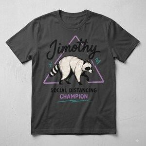 May include: A charcoal grey short-sleeve t-shirt featuring a graphic of a raccoon inside a light purple triangle. Above the raccoon is the text Jimothy in black script. Below the raccoon is the text SOCIAL DISTANCING CHAMPION in black and purple.