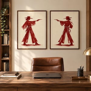 May include: Two framed wall art prints featuring a dark red ink illustration of a figure wearing a wide-brimmed hat, a fringed top, and wide-leg trousers, holding a pistol in an outstretched arm, set against a light cream-colored background.