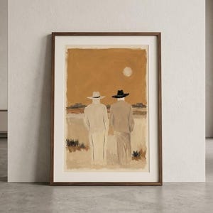 May include: A framed wall art print depicting two figures from behind, wearing wide-brimmed hats and long coats in a desert landscape. The colour palette features warm ochre, beige, and brown tones with a pale sun in the sky above the horizon.