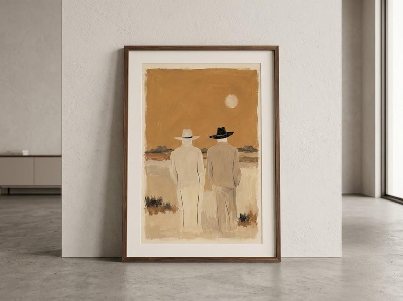 May include: A framed wall art print depicting two figures from behind, wearing wide-brimmed hats and long coats in a desert landscape. The colour palette features warm ochre, beige, and brown tones with a pale sun in the sky above the horizon.
