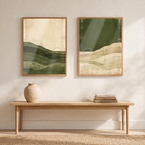 May include: Two rectangular framed wall art prints featuring abstract layered landscapes in shades of olive green, cream, and beige. Each print displays a thin arched line representing a sun. The prints are displayed side by side on a wall.