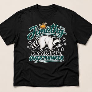 May include: A black short-sleeved t-shirt featuring a graphic of a raccoon wearing a crown. The text reads Jimothy above the raccoon and PROFESSIONAL OVERTHINKER below it, surrounded by small star shapes in a teal and white colour scheme.
