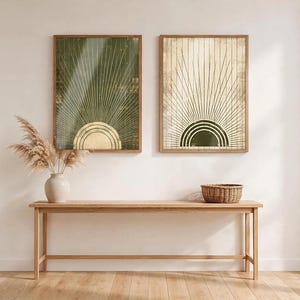 May include: Two rectangular wall art prints in wooden frames featuring a sunburst design. The left print has a dark green background with a gold sun, while the right print has a cream background with a dark green sun. Both display radiating lines.