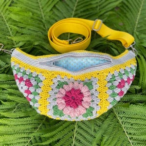 May include: A crescent-shaped crochet fanny pack featuring a colorful granny square pattern with pink, yellow, green, and gray yarn. The bag has a yellow adjustable strap, a gray zipper closure, and a light blue interior lining with a small pattern.