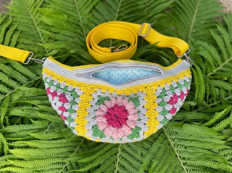 May include: A crescent-shaped crochet fanny pack featuring a colorful granny square pattern with pink, yellow, green, and gray yarn. The bag has a yellow adjustable strap, a gray zipper closure, and a light blue interior lining with a small pattern.