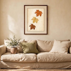 May include: A framed wall art print featuring three maple leaves in shades of orange, yellow, and brown arranged vertically on a light, textured background. The artwork is enclosed in a simple dark brown wooden frame with a white mat border.