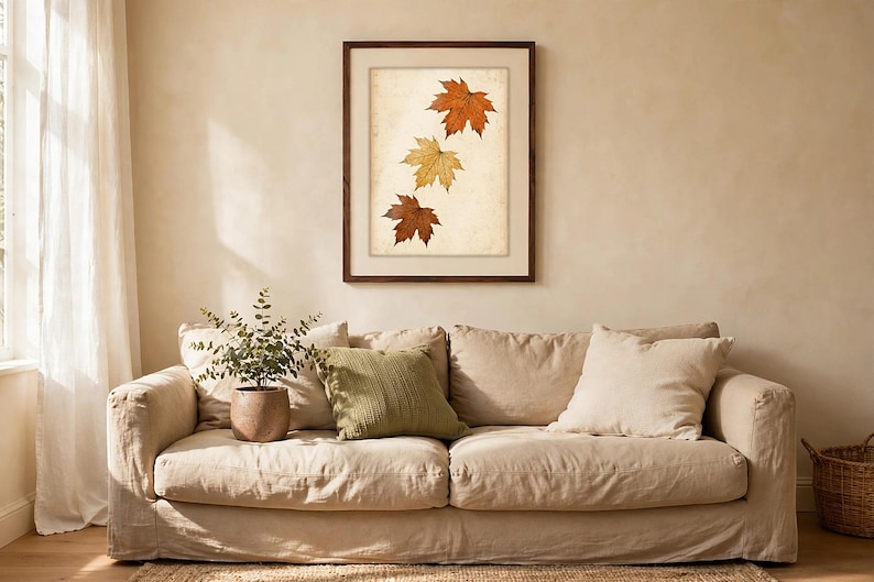 May include: A framed wall art print featuring three maple leaves in shades of orange, yellow, and brown arranged vertically on a light, textured background. The artwork is enclosed in a simple dark brown wooden frame with a white mat border.