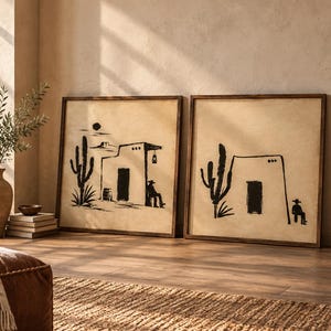 May include: Two square framed wall art pieces featuring black ink illustrations on a beige background. The artwork depicts desert scenes with saguaro cacti, adobe-style buildings, and a figure wearing a cowboy hat sitting near the structures.