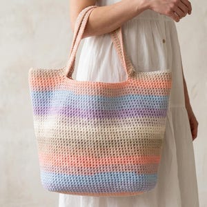 May include: A large, rectangular crochet tote bag featuring horizontal stripes in a gradient of soft pastel colors including peach, light blue, lavender, cream, and tan. The bag has two integrated shoulder straps and a textured, woven stitch pattern.
