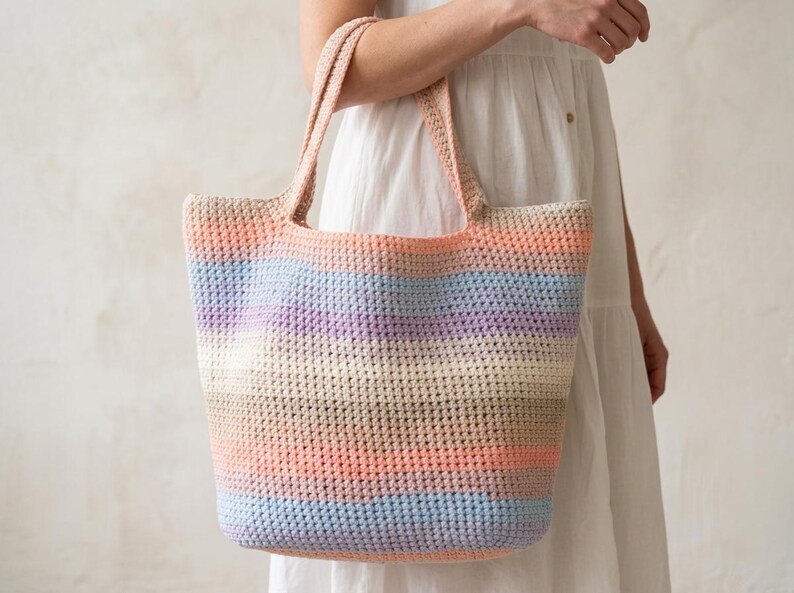 May include: A large, rectangular crochet tote bag featuring horizontal stripes in a gradient of soft pastel colors including peach, light blue, lavender, cream, and tan. The bag has two integrated shoulder straps and a textured, woven stitch pattern.