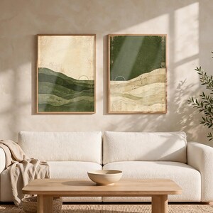 May include: Two framed rectangular wall art prints featuring abstract landscape designs. The left print displays layered green hills against a cream background, while the right print shows a dark green sky over beige rolling hills and a sun icon.
