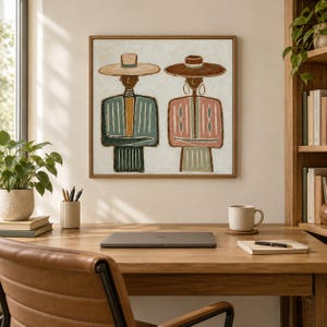 May include: A square framed wall art piece featuring a stylized painting of two figures wearing wide-brimmed hats. The left figure wears a blue and tan striped garment, while the right figure wears a pink and tan striped garment against a cream background.