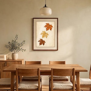 May include: A framed wall art print featuring three maple leaves in shades of orange, yellow, and brown arranged vertically on a light, textured background. The artwork is enclosed in a simple dark brown wooden frame.