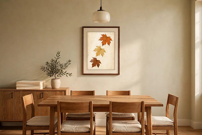 May include: A framed wall art print featuring three maple leaves in shades of orange, yellow, and brown arranged vertically on a light, textured background. The artwork is enclosed in a simple dark brown wooden frame.