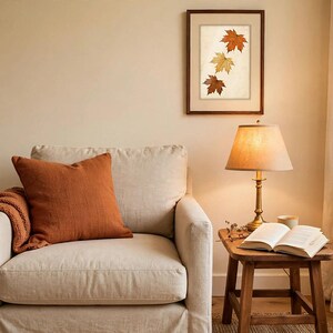 May include: A beige upholstered armchair features a burnt orange throw pillow and a matching textured blanket. Beside it, a wooden side table holds a brass lamp, an open book, and a small cup. A framed print of three autumn leaves hangs above.