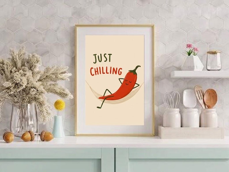 May include: A framed wall art print featuring a cartoon red chili pepper with a face, arms, and legs, relaxing in a hammock. The text "JUST CHILLING" is printed in dark green and red above the chili pepper on a light cream background.