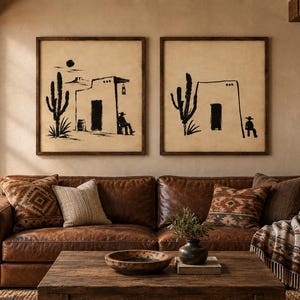 May include: Two square framed wall art pieces featuring black ink illustrations of desert scenes with cacti, adobe-style buildings, and figures wearing hats, set against a light beige background, displayed above a brown leather sofa.