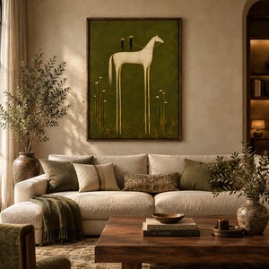 May include: A framed wall art print featuring a stylized white horse with elongated legs standing on a dark olive green background. Two small figures in hats are perched on the horse's back, surrounded by thin, vertical white floral stems.